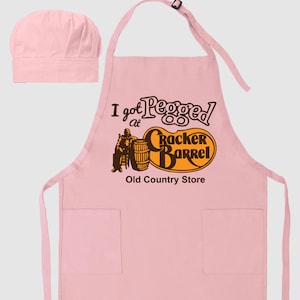 May include: A light pink apron and chef's hat set. The apron features the text "I got Pegged at Cracker Barrel Old Country Store" in brown and orange. The apron has two pockets and an adjustable neck strap.
