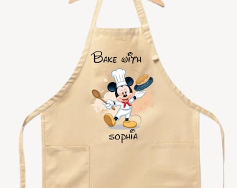 May include: Tan apron featuring a cartoon Mickey Mouse chef holding a wooden spoon and a pie. The apron has the text "Bake with" above the character and the name "Sophia" below. Includes two front pockets.