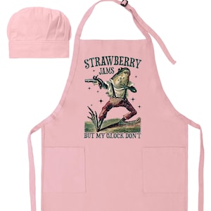 May include: A light pink apron and chef's hat set. The apron features a frog illustration holding a gun, with the text "STRAWBERRY JAMS" above and "BUT MY GLOCK DON'T" below. The chef's hat is a matching light pink color.