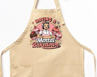 Könnte beinhalten: Eine beige Schürze mit einer Tasche auf der Vorderseite. Die Schürze zeigt ein Grafikmotiv einer Frau mit einem Nudelholz und Cupcakes mit dem Text "Having a Mental Bakedown".