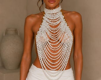 Könnte beinhalten: Ein Neckholder-Oberteil aus mehreren kaskadierenden Strängen weißer Kunstperlen. Das Design verfügt über einen hohen, chokerartigen Kragen aus Perlenreihen, wobei zusätzliche drapierte Perlenlagen über den Oberkörper verlaufen, die an den Seiten durch dünne goldfarbene Metallketten verbunden sind.