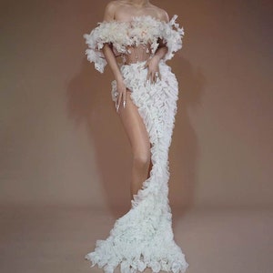 May include: A white off-the-shoulder gown with a ruffled skirt and a fitted bodice. The gown is made of a sheer fabric with a delicate floral pattern.