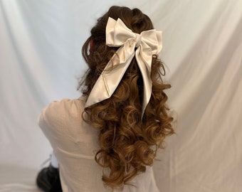 May include: A white satin bow hair clip with a large bow and long tails. The bow is tied in a simple knot and the tails are flowing down the back of the head.