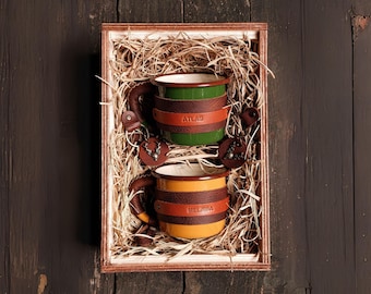 May include: Two enamel mugs with leather handles and personalized names, "Atlas" and "Melissa", are nestled in a wooden box filled with straw.