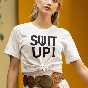 May include: A white t-shirt with the text "Suit Up!" printed in black. The shirt is tied at the waist and worn with a white tiered maxi skirt and a brown leather belt with metal accents.