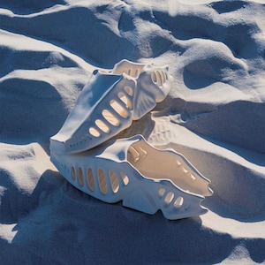 May include: Two white, open-toe sandals with a unique, geometric design. The sandals have a series of oval cutouts along the sides and top, and are resting on a bed of white sand. The sandals appear to be made of a hard material.