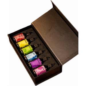 May include: A black box with a lid open, revealing six small glass bottles with colorful labels. The labels feature different botanical illustrations and names, including "Chaga", "Goji", "Green Tea", "Reishi", "Lion's Mane", and "Shiitake".