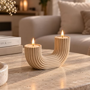 Peut inclure: Un bougeoir en forme de U fabriqué dans un matériau beige avec une texture nervurée verticale. Le support présente deux ouvertures circulaires sur le dessus, chacune contenant une bougie chauffe-plat allumée avec une flamme visible.