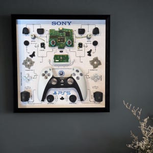 Könnte beinhalten: Ein gerahmter weißer und schwarzer PlayStation 5-Controller mit dem Aufdruck "PS5". Der Rahmen enthält auch die einzelnen Teile des Controllers, darunter die Leiterplatten, Tasten und Gummipads.