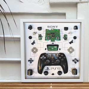 May include: A white framed artwork featuring a disassembled PlayStation 5 controller. The controller is laid out on a white background with each component labeled. The artwork includes the words "SONY" and "PS5".