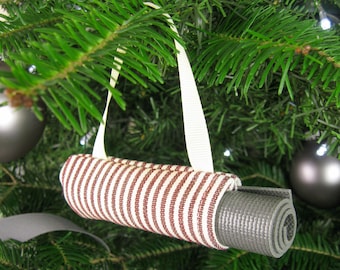 May include: A Christmas tree ornament shaped like a rolled-up yoga mat. The mat is gray and is held in place by a fabric tube with red and white stripes. A white ribbon is used to hang the ornament on the tree.