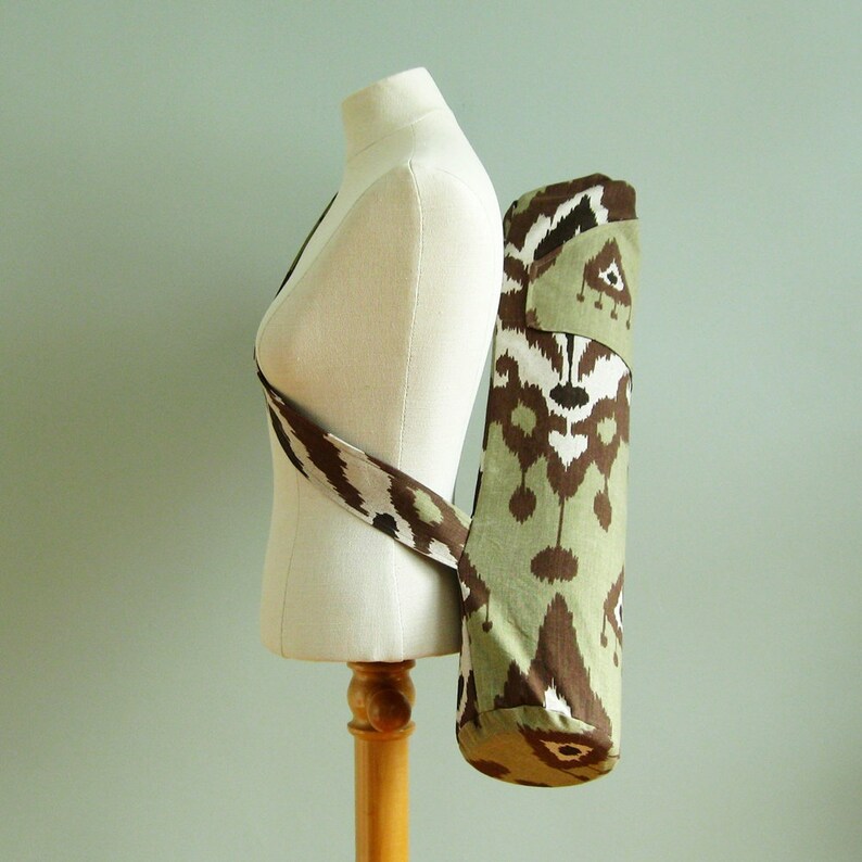 May include: A brown and green yoga mat bag with a patterned design. The bag has a long strap that can be worn over the shoulder.