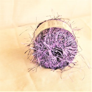 May include: A ball of yarn with a mix of purple, black, and white fibers. The yarn has a textured, fringed appearance. A label is wrapped around the center of the ball. The yarn is likely used for knitting or crocheting projects.