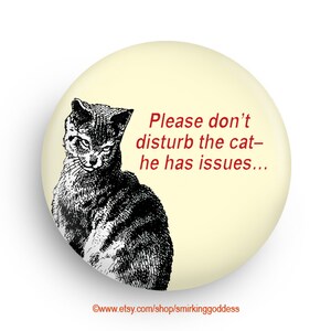 May include: A round button with a yellow background features a black and white illustration of a cat with a grumpy expression. The text "Please don't disturb the cat - he has issues..." is written in red.