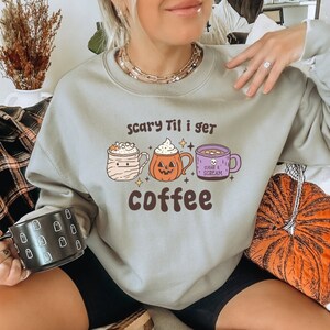 May include: A light gray sweatshirt with the text "scary Til I get coffee" and three cartoon coffee mugs. The mugs are designed as a mummy, a jack-o'-lantern, and a purple mug with a skull. The sweatshirt is a Halloween-themed design.