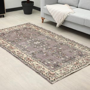 Puede incluir: Una alfombra rectangular de estilo vintage con un campo central morado apagado y un borde floral detallado en crema, rojo y verde. La alfombra está colocada sobre un suelo de madera clara, cerca de un sofá gris.