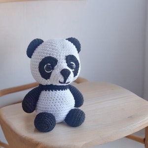 May include: A crocheted panda bear plush toy with black ears, eyes, nose, and paws. The panda is sitting on a wooden chair.