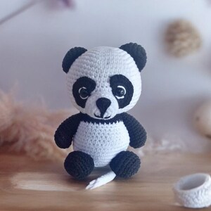 May include: A crocheted panda bear plush toy with black eyes and a white body. The panda is sitting on a wooden surface.