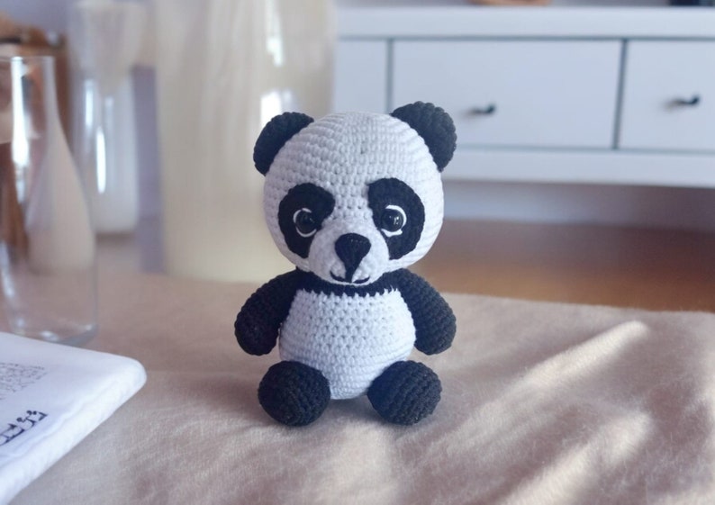 May include: A small crocheted panda bear toy with black and white fur. The panda has black eyes and a black nose.