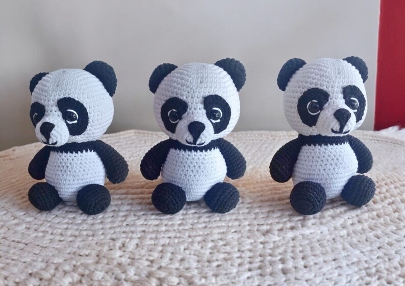 May include: Three crocheted panda bears, each with black eyes, a black nose, and black ears. The pandas are sitting on a white crocheted surface.