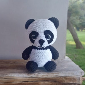 May include: A white crocheted panda bear with black eyes, ears, and paws. The panda is sitting on a wooden surface.
