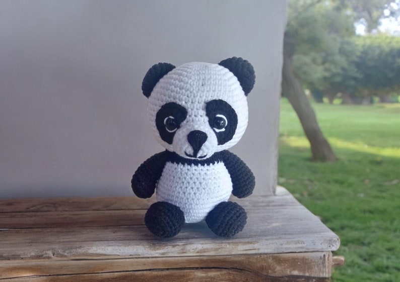 May include: A white crocheted panda bear with black eyes, ears, and paws. The panda is sitting on a wooden surface.