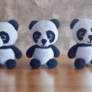 May include: Three crocheted panda bears, each with black eyes, a black nose, and black ears. The pandas are sitting on a wooden surface.