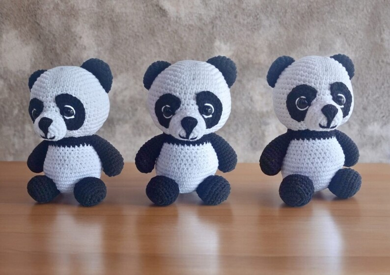 May include: Three crocheted panda bears, each with black eyes, a black nose, and black ears. The pandas are sitting on a wooden surface.