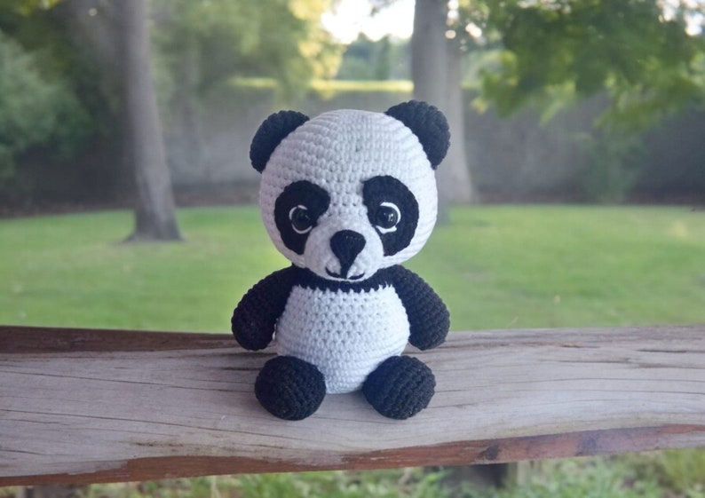May include: A crocheted panda bear plush toy with black and white fur. The panda is sitting on a wooden surface.