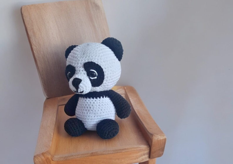 May include: A crocheted panda bear plush toy with black ears, eyes, and paws, sitting on a wooden chair.