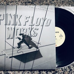 May include: A monochrome album cover for Pink Floyd's "Works" LP. The cover displays the band's name in bold lettering, with an illustration of a person running on a metallic structure. A vinyl record is partially visible.