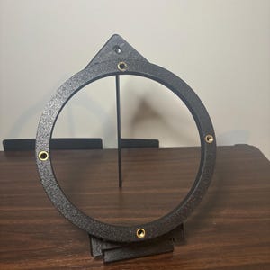 May include: A black circular frame with a textured finish, featuring four evenly spaced brass-colored metal inserts. The frame includes a triangular mounting tab at the top and is supported by a vertical stand attached to a rectangular base.