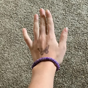 Op de afbeelding: Een elastisch haarbandje met paarse en witte strepen om de pols van iemand.