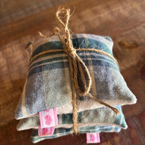 May include: A stack of three square, plaid fabric sachets tied with twine. The sachets are in shades of blue, green, and beige. Pink ribbon tags with a heart design are attached to the sachets. The twine is knotted at the top.