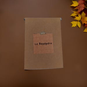 Peut inclure: Un carnet brun avec une note en papier beige fixée sur le devant. La note porte le texte "Le Repêpère" écrit en noir.