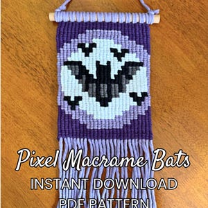 Può includere: Un arazzo in macramè con un design a pipistrello pixelato nei toni del viola, viola chiaro, nero e bianco, appeso a un bastoncino di legno con un cordino. Il testo recita: Pixel Macrame Bats INSTANT DOWNLOAD PDF PATTERN.