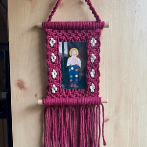 May include: A handmade macrame wall hanging in a deep magenta color. The frame surrounds a small picture of a doll. White flower accents are placed around the picture. The bottom of the frame has long fringe.