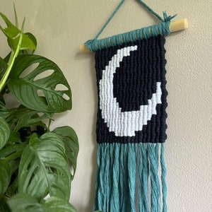 Peut inclure: Une tapisserie murale tissée en noir et blanc avec un motif de croissant de lune. La tapisserie a des franges turquoise et est fixée à un bâtonnet en bois.