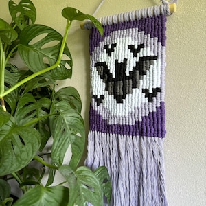 May include: A purple and white macrame wall hanging with a black bat design. The hanging has a fringe border.