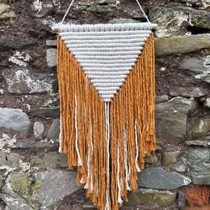 May include: A macramé wall hanging with a triangular design. The top section is grey, and the bottom features long, orange and white fringe. It is suspended from a wooden dowel with a white string.