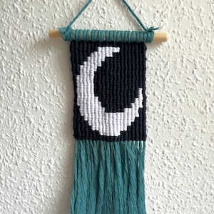 May include: A macrame wall hanging with a crescent moon design. It has a navy blue background with a white moon, teal fringe, and a wooden dowel. The hanging is approximately 30 cm long.
