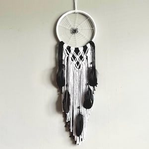 Peut inclure: Un attrape-rêves blanc et noir avec un motif de toile d'araignée et des plumes noires qui pendent du bas.
