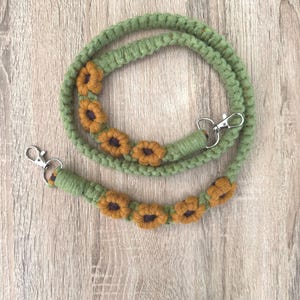 May include: A green macrame lanyard with a sunflower design. The lanyard features small, yellow sunflowers with brown centres. The lanyard has silver-coloured metal clasps on each end.