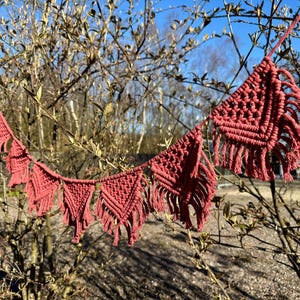 Peut inclure: Guirlande macramé faite à la main, de couleur rose poudré. Elle présente des formes triangulaires et carrées avec des nœuds complexes et des franges. La guirlande est suspendue à l'extérieur, devant un arrière-plan flou d'arbres et de ciel bleu.