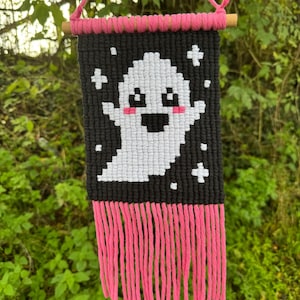 Puede incluir: Un colgante de pared de macram&eacute; hecho a mano con un fantasma blanco pixelado sobre un fondo negro. Flecos y acentos rosas a&ntilde;aden color. El fantasma tiene mejillas rosadas y una boca sonriente. Se utiliza una clavija de madera y un cord&oacute;n rosa para colgar.