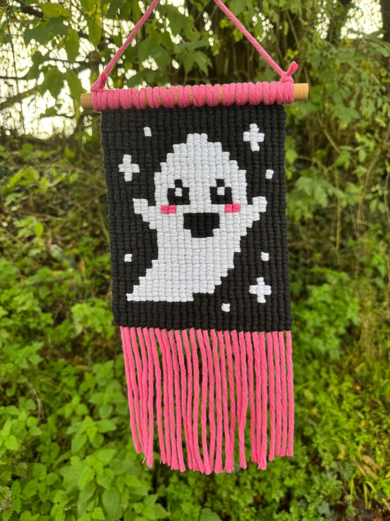 Puede incluir: Un colgante de pared de macram&eacute; hecho a mano con un fantasma blanco pixelado sobre un fondo negro. Flecos y acentos rosas a&ntilde;aden color. El fantasma tiene mejillas rosadas y una boca sonriente. Se utiliza una clavija de madera y un cord&oacute;n rosa para colgar.