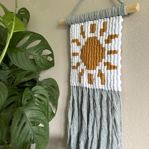 May include: A wall hanging with a woven sun design in shades of white and mustard yellow. The sun is surrounded by a white border and the hanging has a fringe of light blue yarn.