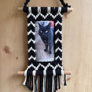 May include: A black and white macrame wall hanging with a photo of a black cat. The frame is made of black and white knotted rope, with a wooden dowel at the top and bottom. The hanging has a fringe at the bottom.