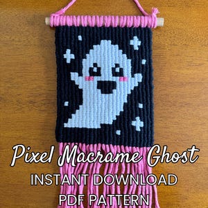 May include: A macrame wall hanging featuring a pixelated ghost design in light blue and white against a black background, accented with pink cheeks. The piece is suspended from a wooden dowel by a pink cord and finished with pink fringe.