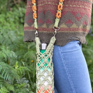 May include: A green, white and orange crocheted bottle holder with a silver clip. The holder is attached to a long orange and green crocheted strap.